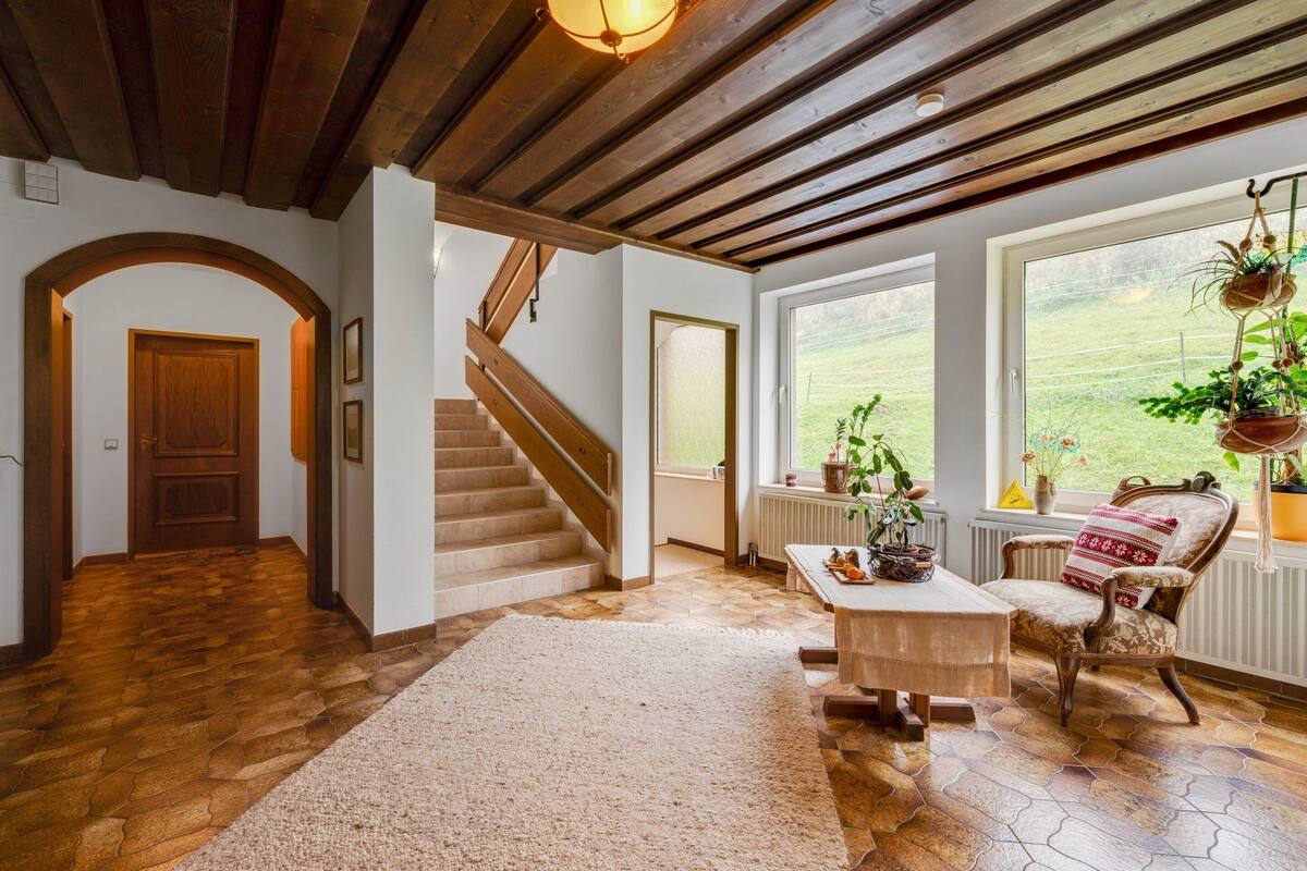 Un vaste espace de vie agrémenté d’un escalier en bois, d’une grande fenêtre offrant une vue sur l’extérieur et d’un coin salon confortable avec un fauteuil à motifs et une petite table.