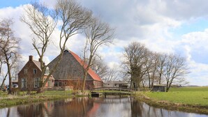 A traditional brick house with a red roof, a small bridge over a calm water body, and a grassy field in the background. - Secluded Chalet in Suwald With Hottub (Suwâld)