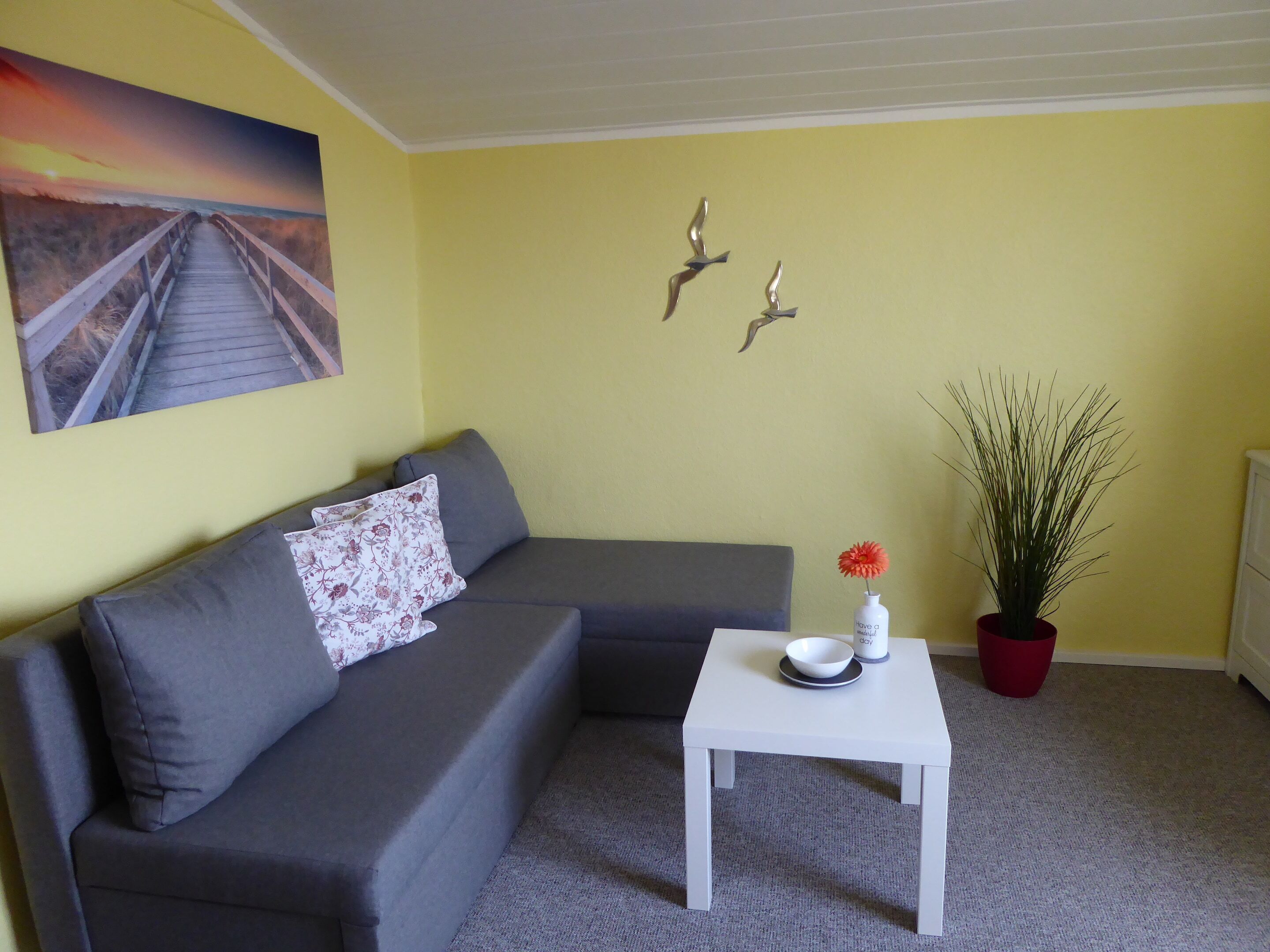 A living room with a grey sofa, a white coffee table, a red potted plant, and a painting of a sunset over a beach.
