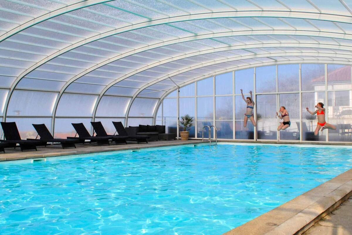 An indoor swimming pool with clear glass walls, lounge chairs, and a pool ladder.