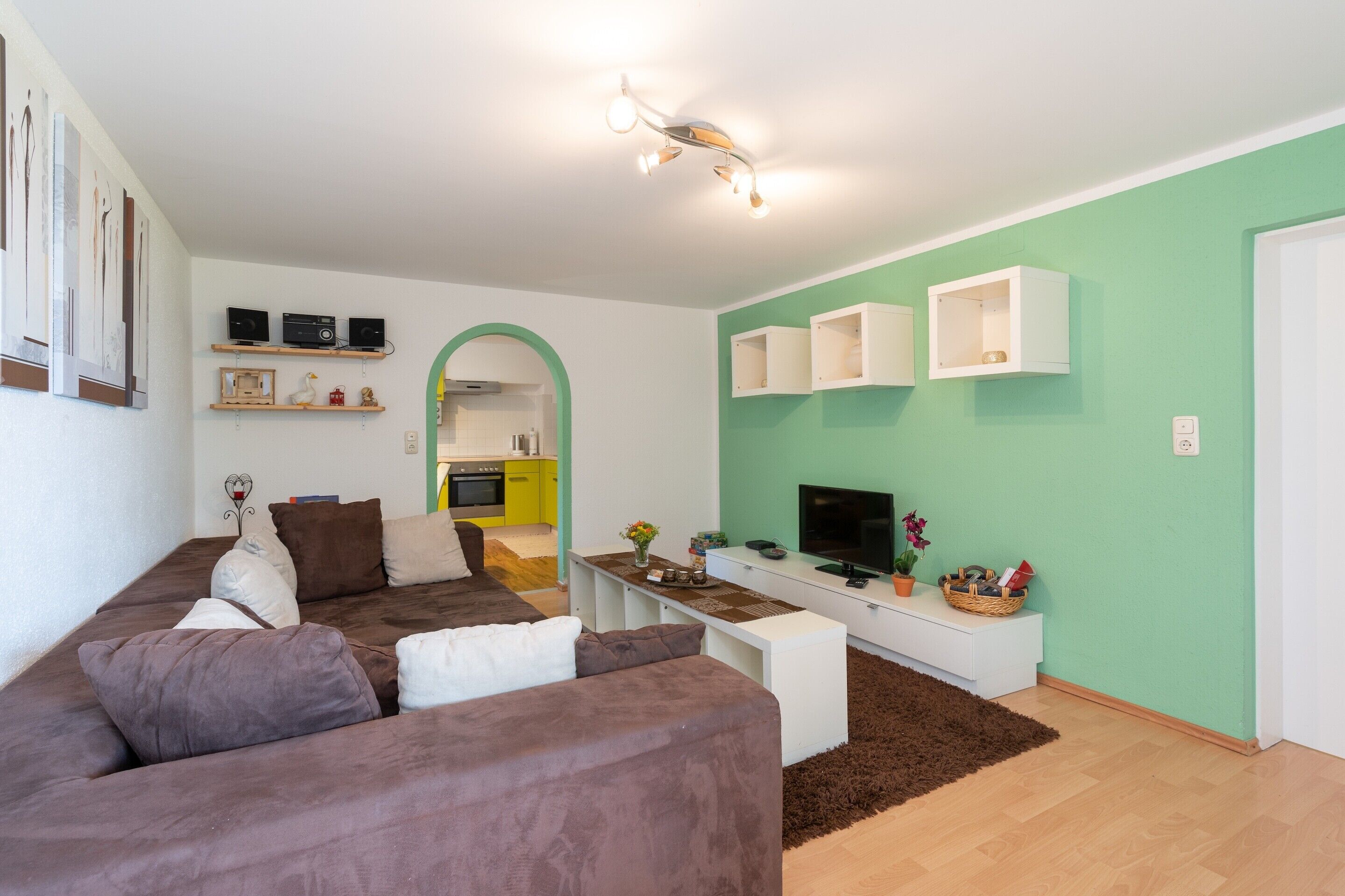 A modern living room with a brown sofa, a white coffee table, and a TV on a white stand. There is a kitchen visible through an archway, and a green wall with white cabinets.