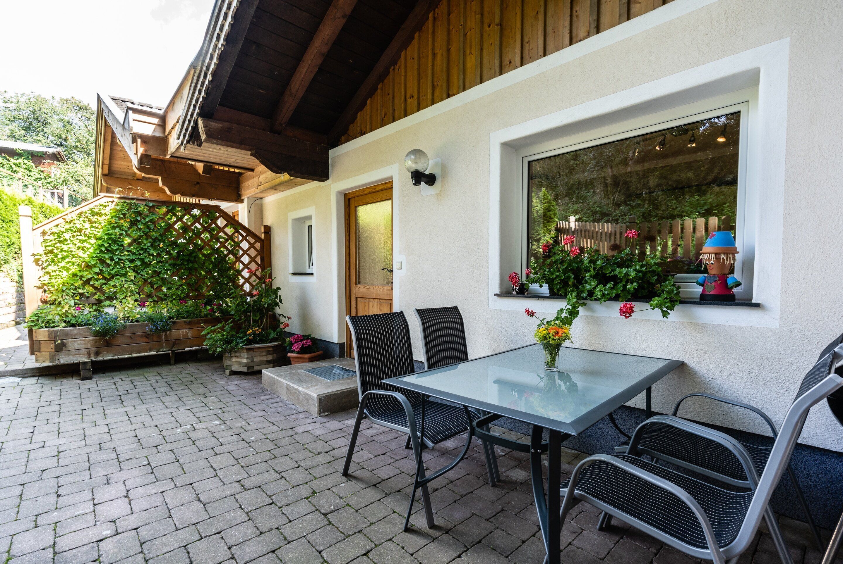 An outdoor patio with a glass table and metal chairs, surrounded by potted plants and a wooden trellis.