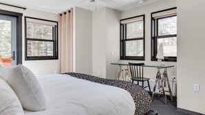 A bedroom with a bed, a desk, and a chair. There are windows with curtains, a ceiling fan, and a radiator.