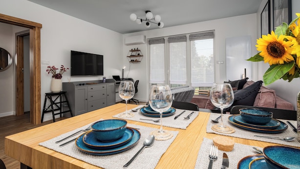 A dining area with a wooden table set for four, a kitchen in the background, and a large window with curtains. - Apartamenty Sun & Snow Nad Ujściem (Leba)
