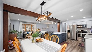 Une cuisine moderne avec des meubles blancs, des appareils en acier inoxydable et un coin repas agrémenté d’une nappe à rayures et de chaises en bois.
