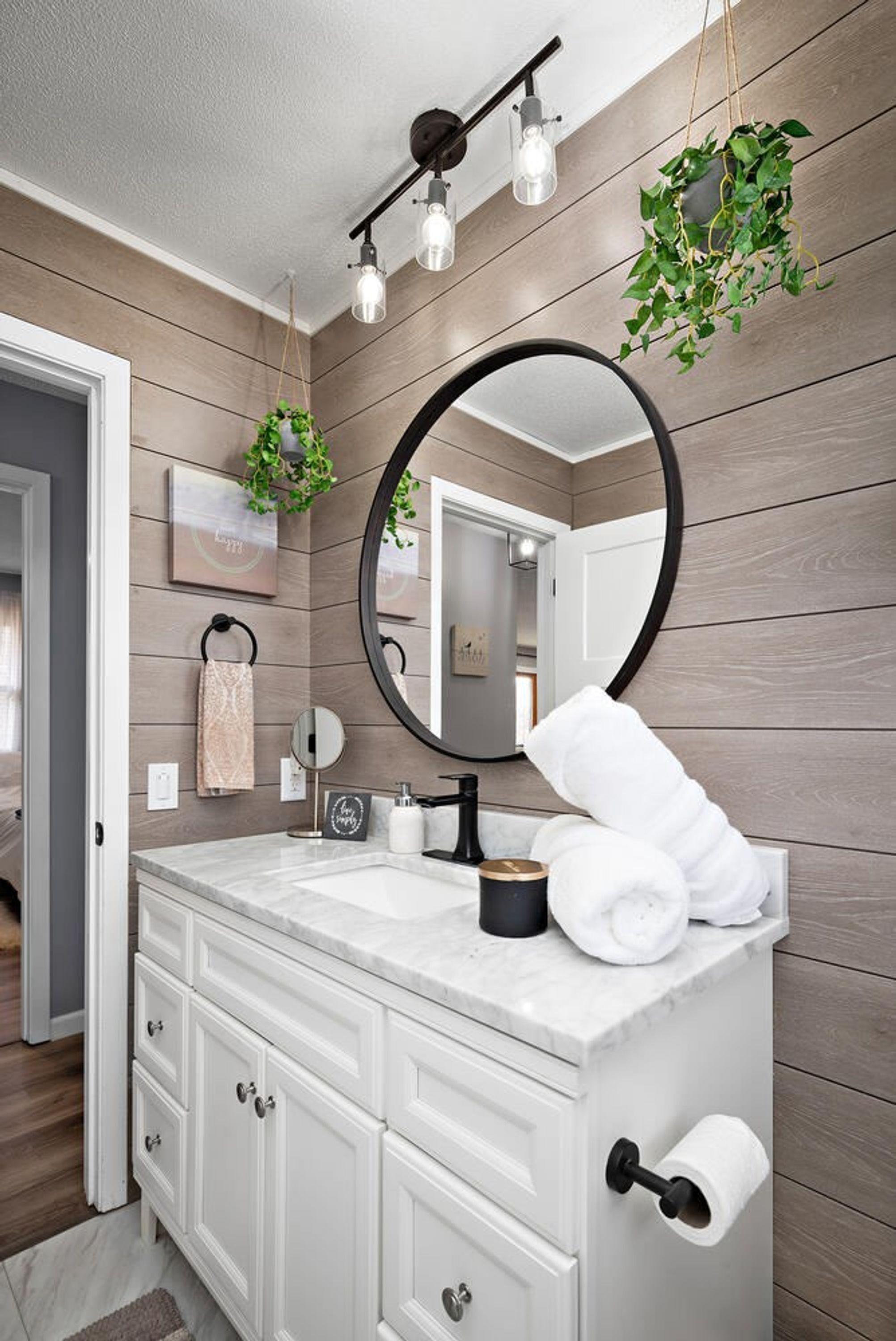 A bathroom with a wooden wall, a white vanity, a round mirror, a towel rack, and a hanging plant.