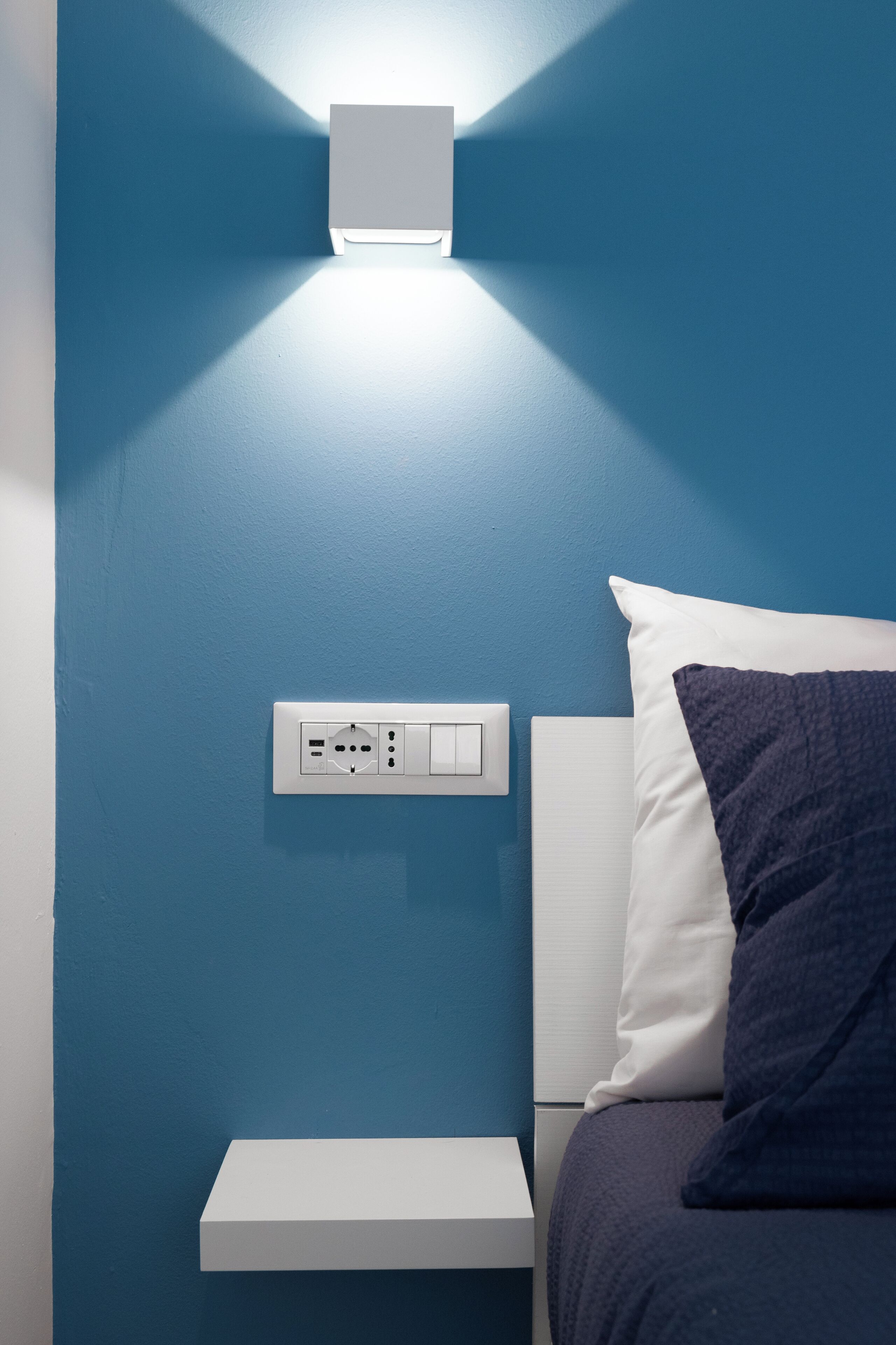 Una camera da letto con una parete blu, un comodino, un letto con cuscino bianco e blu scuro, e un punto luce fissato alla parete.