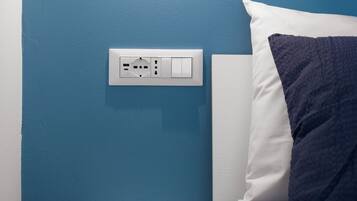 Una camera da letto con una parete blu, un comodino, un letto con cuscino bianco e blu scuro, e un punto luce fissato alla parete.