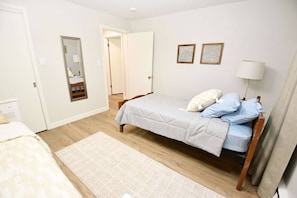 A bedroom with a bed, a bedside table, a lamp, a mirror, and a door leading to another room.
