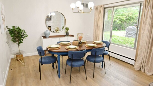 A dining room with a round table set for six, blue chairs, a mirror, a chandelier, and a view of greenery outside. - Mid Century Modern, 10 min walk to ND! (1831) (South Bend)
