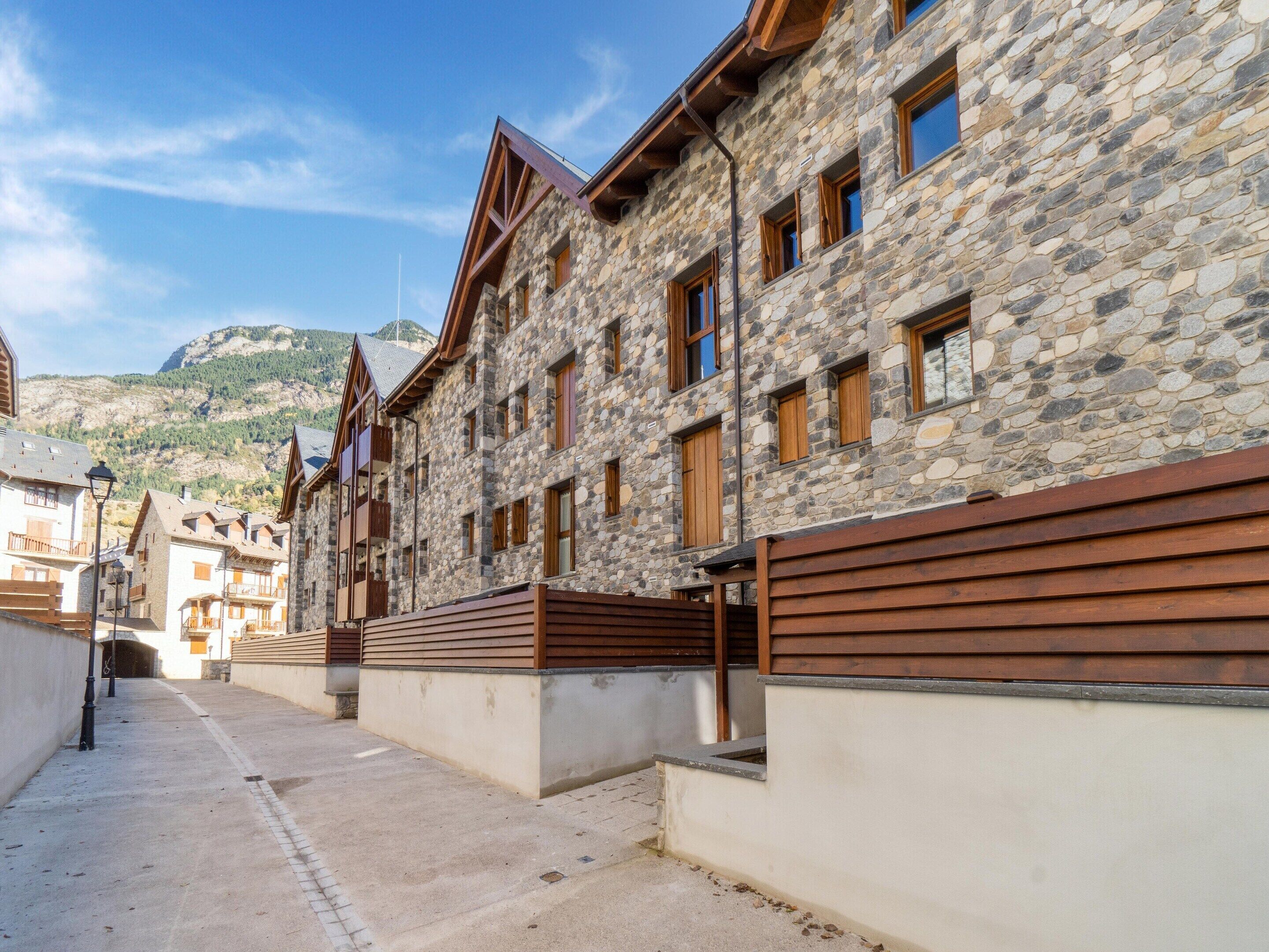 A stone building with multiple windows and balconies, a concrete pathway, and a mountainous backdrop.