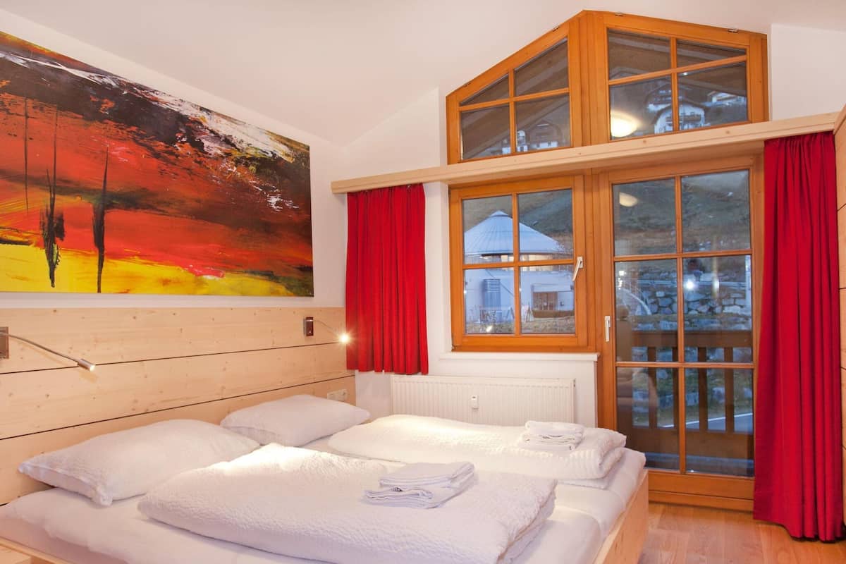 A room with a wooden headboard, a large painting, a window with red curtains, and a view of a house outside.