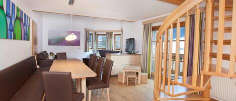 A modern living room with a wooden staircase, a dining area with a wooden table and chairs, and a television on a wooden cabinet.