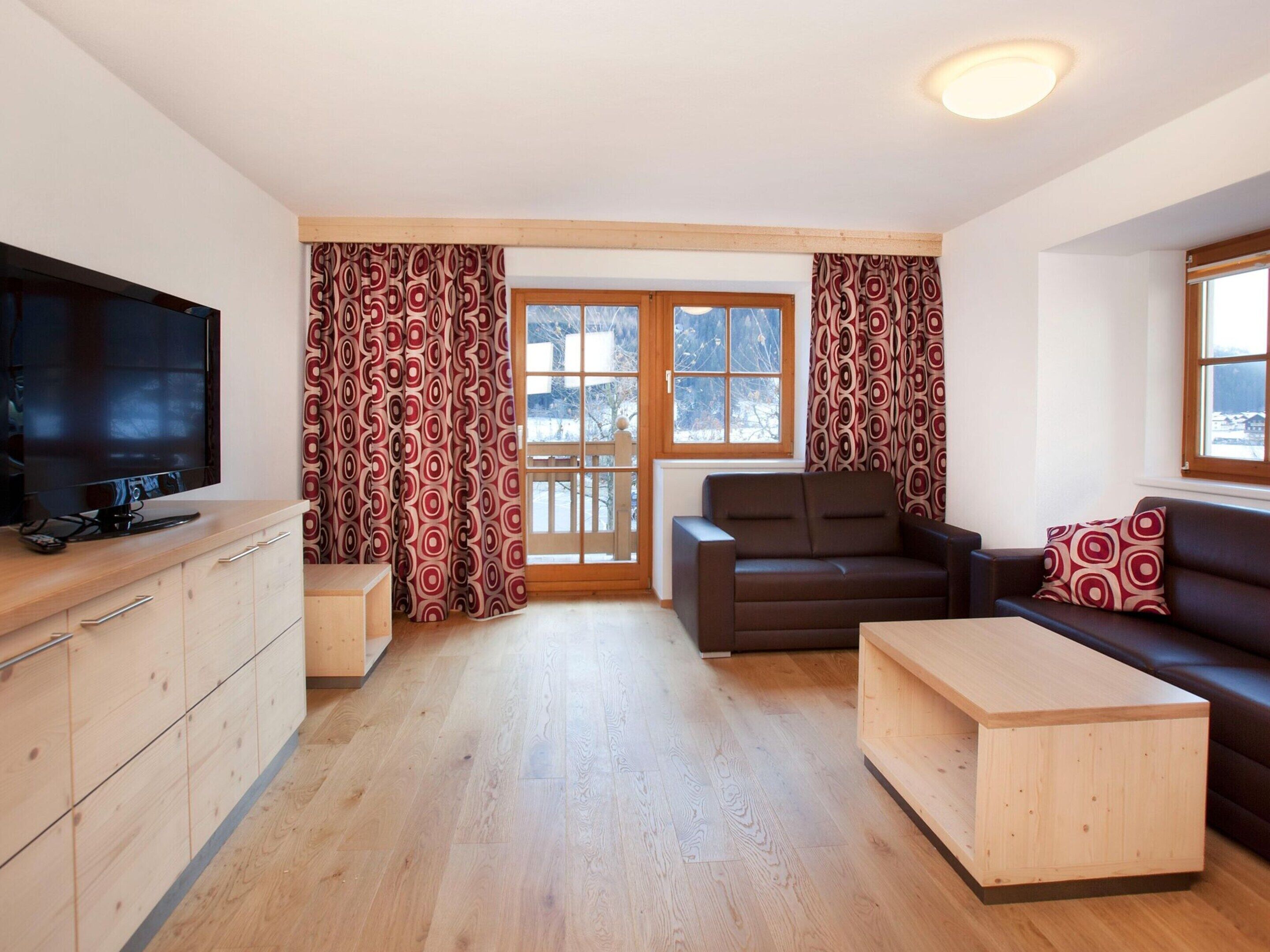 Ein modernes Wohnzimmer mit Flachbildfernseher, Holzmöbeln und gemusterten Vorhängen.