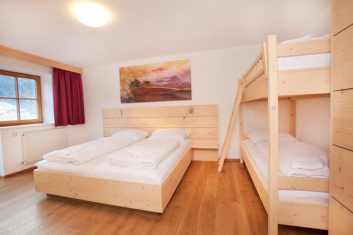 A room with a wooden bunk bed, a single bed, a painting on the wall, and a window with curtains.
