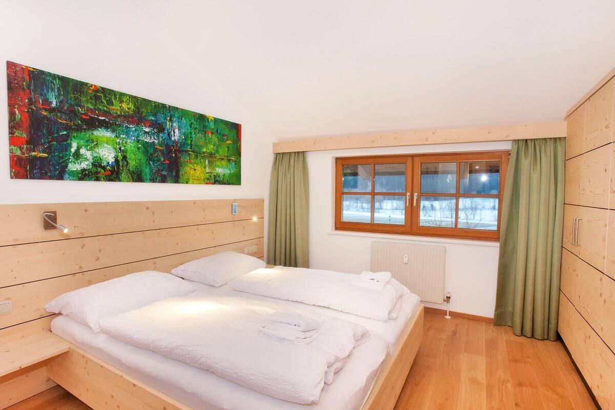 A bedroom with a wooden bed, a window with curtains, a radiator, and a colorful abstract painting on the wall.