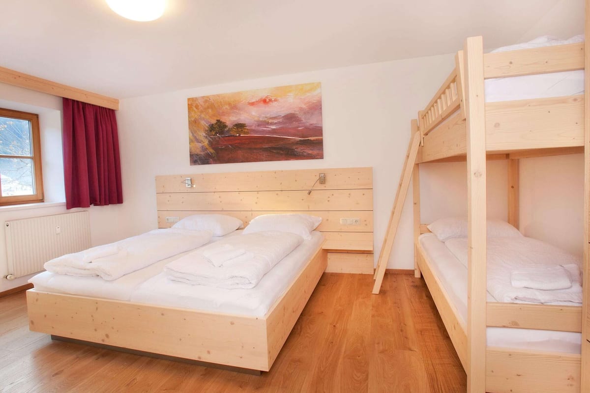 A room with a wooden bunk bed, a single bed, a radiator, a window with curtains, and a painting on the wall.