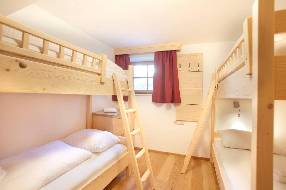 A bunk bed room with a wooden ladder, a window with curtains, and a wooden wardrobe.