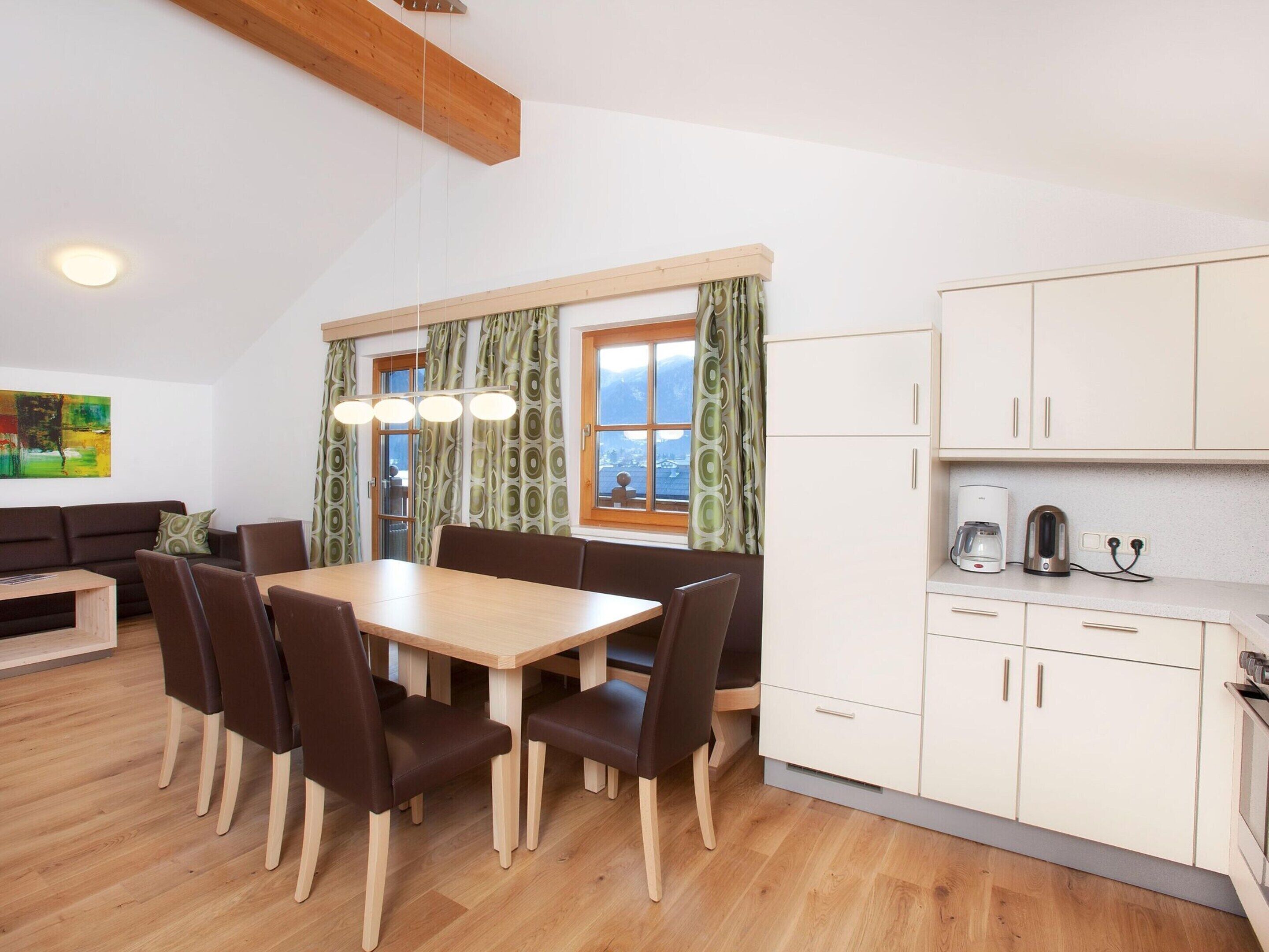 A modern kitchen with white cabinets, a dining table with chairs, and a view of the outdoors through a window with curtains.