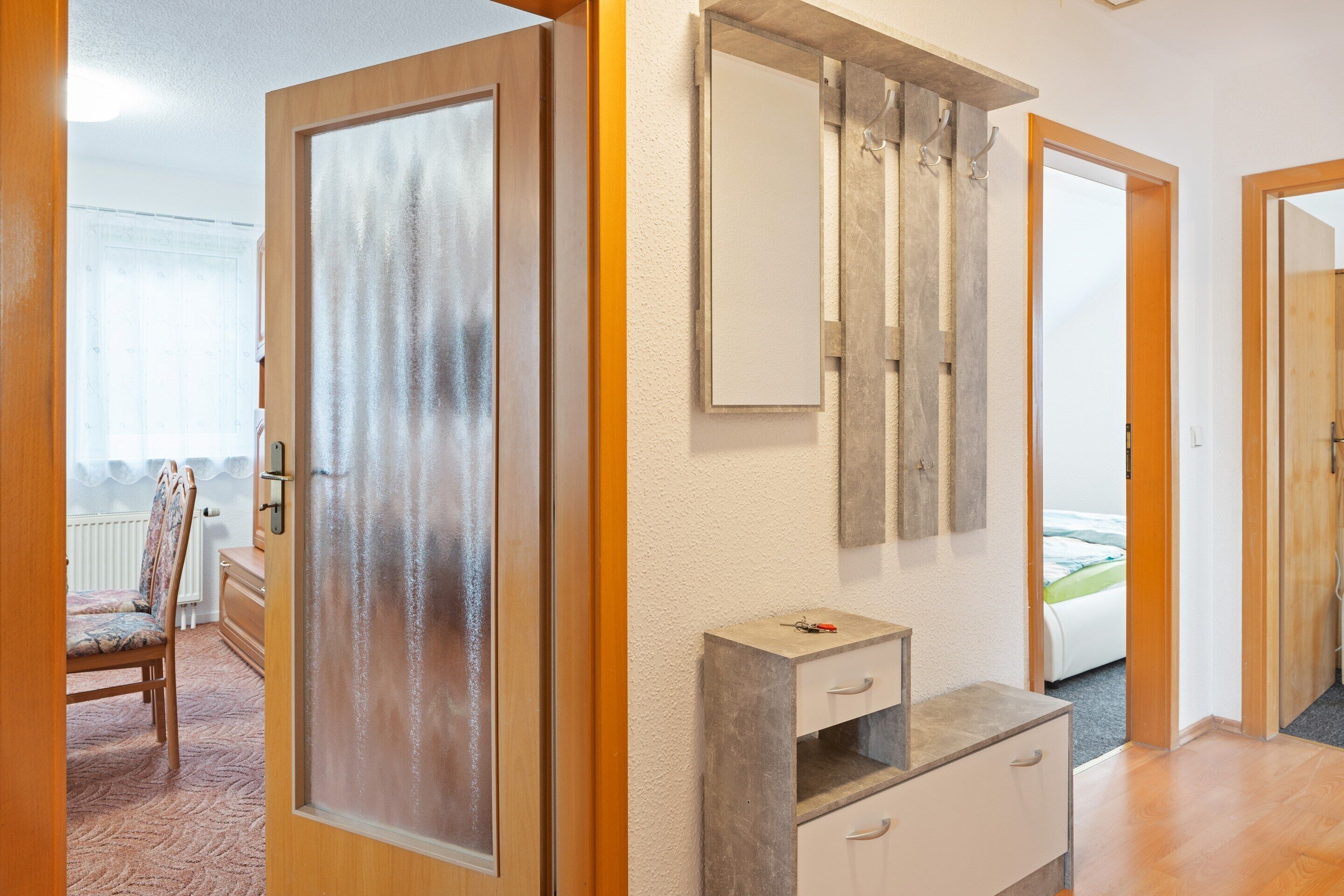 Ein Zimmer mit Holztür, einem Spiegel, einer kleinen Kommode und einem Bett im Hintergrund.