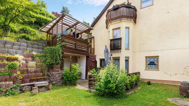 Een huis met twee verdiepingen, een houten terras, een stenen muur en een tuin.