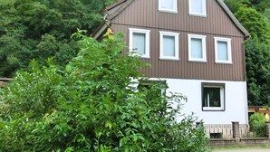 Une maison à deux étages, avec un toit brun et des murs blancs, entourée d’arbres verdoyants et touffus.