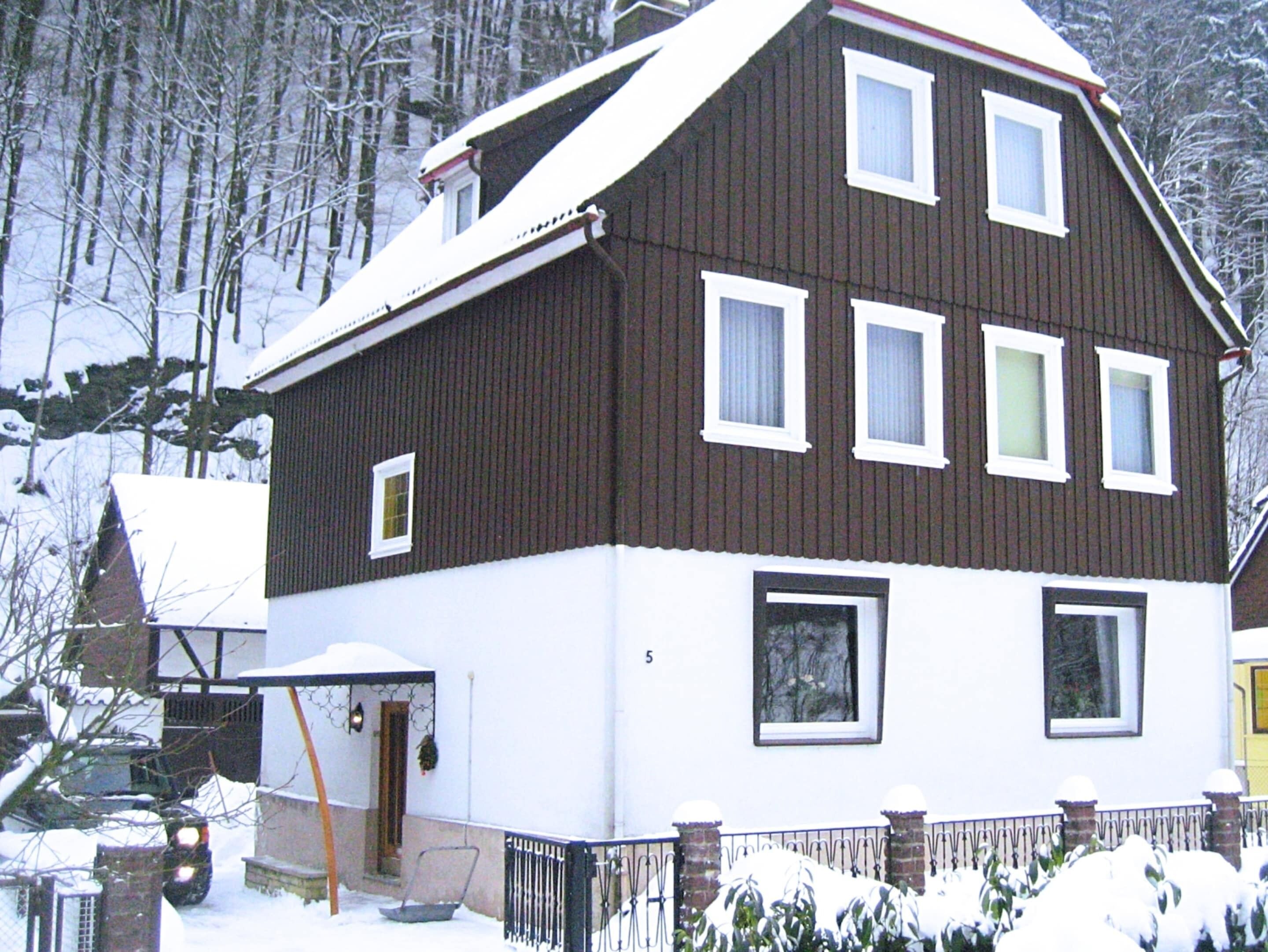 Une maison à deux étages, à l’extérieur brun et aux ornements blancs, entourée d’arbres couverts de neige.