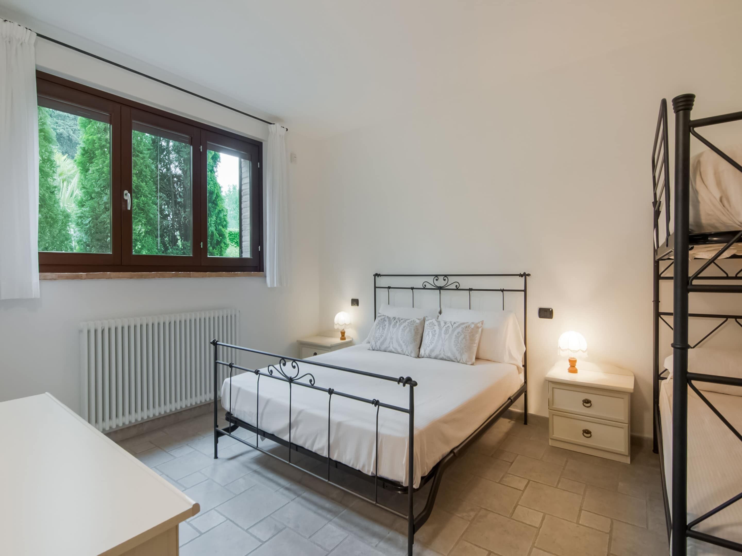 A bedroom with a metal bed frame, a bedside table with a lamp, a radiator, and a window with curtains.