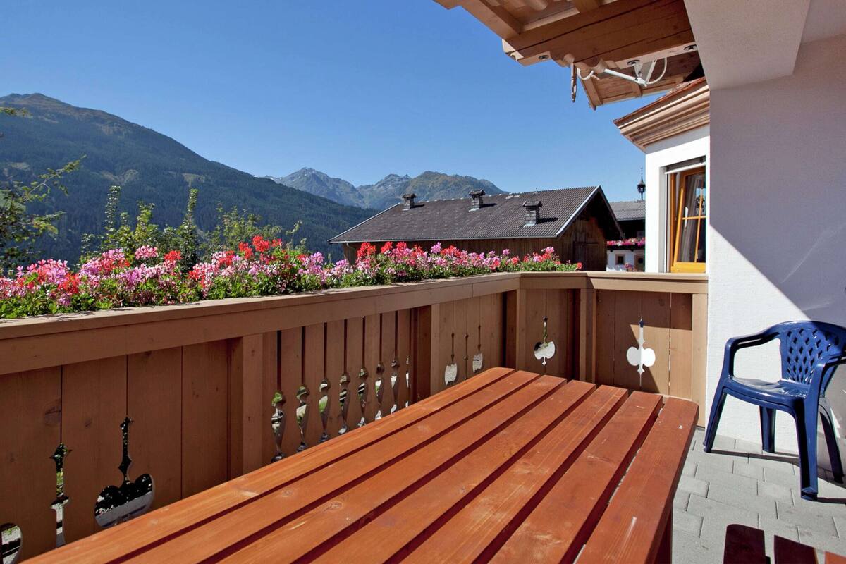 A wooden deck with a bench and a plastic chair, overlooking a mountain range and a building with a sloped roof.