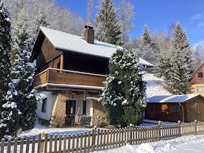 A wooden chalet with a stone foundation, a balcony, and a wooden fence in a snowy landscape.