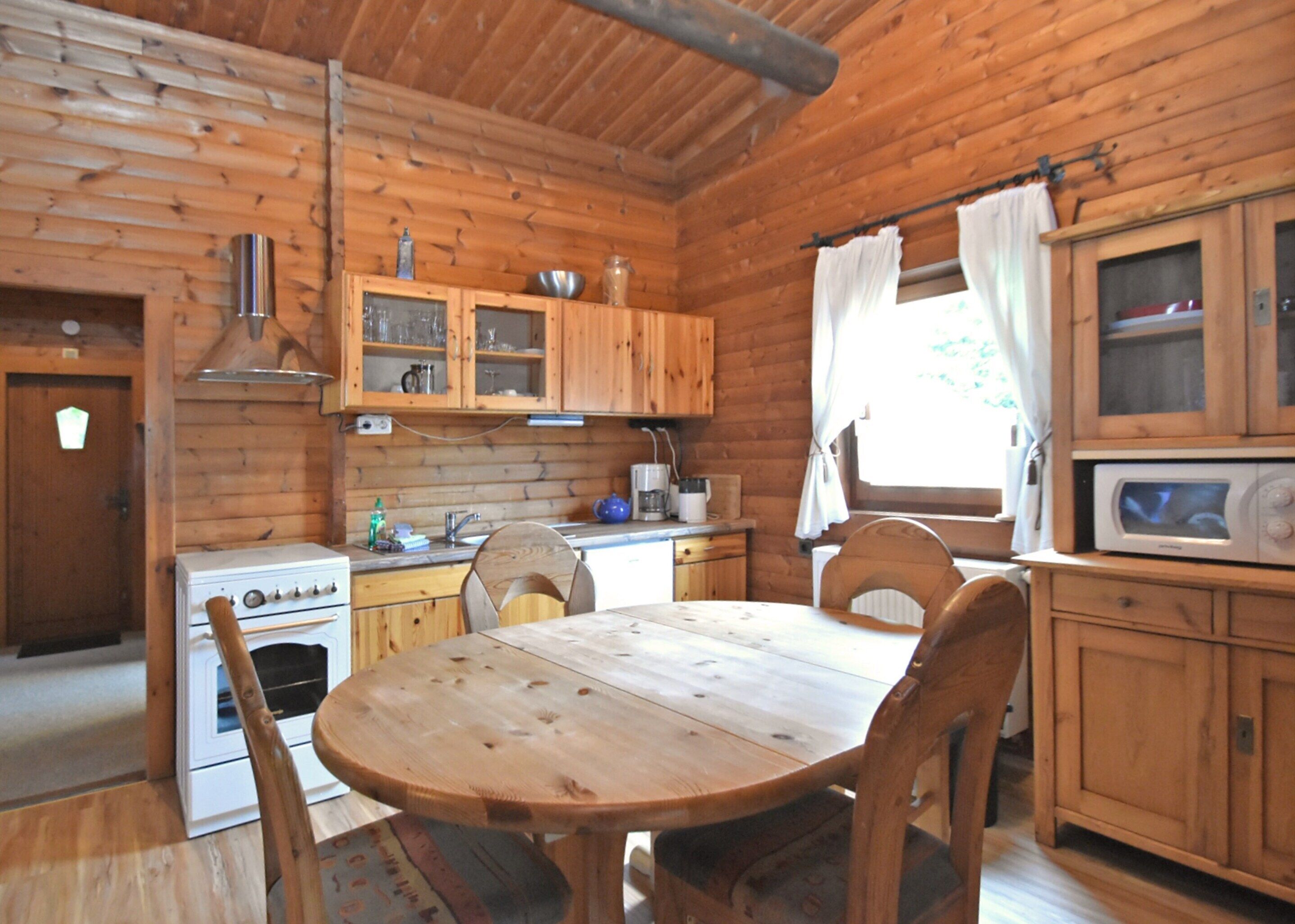 Cocina de una cabaña de madera con una mesa redonda, sillas, estufa, armarios y una ventana con cortinas.
