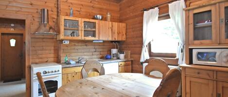 Cocina de una cabaña de madera con una mesa redonda, sillas, estufa, armarios y una ventana con cortinas.