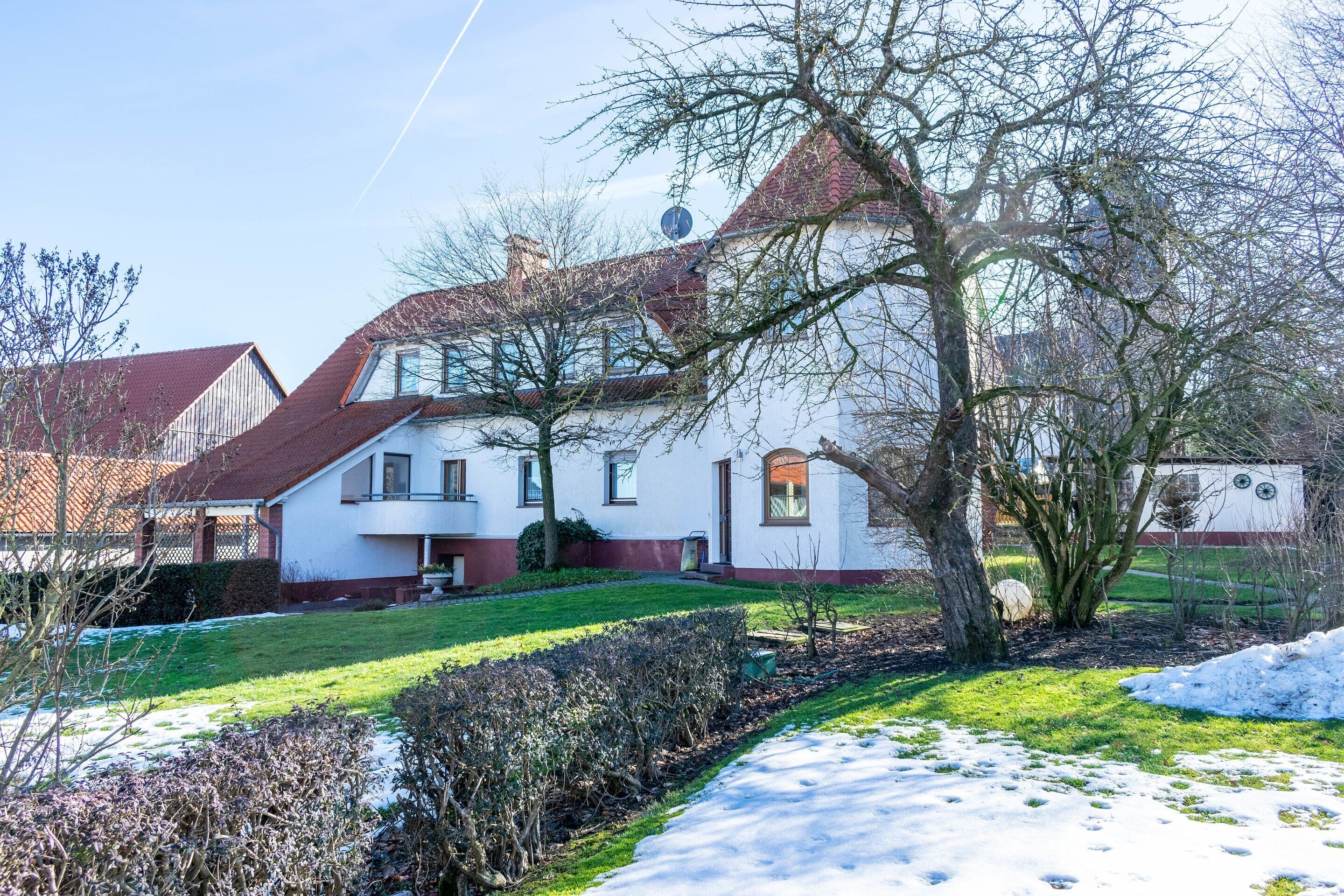 Une maison blanche avec un toit rouge, entourée d’une pelouse enneigée et d’un petit jardin.