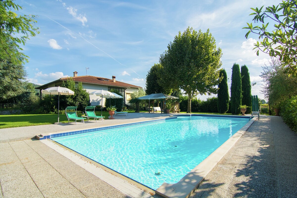 A swimming pool with lounge chairs and umbrellas, surrounded by trees and a house.