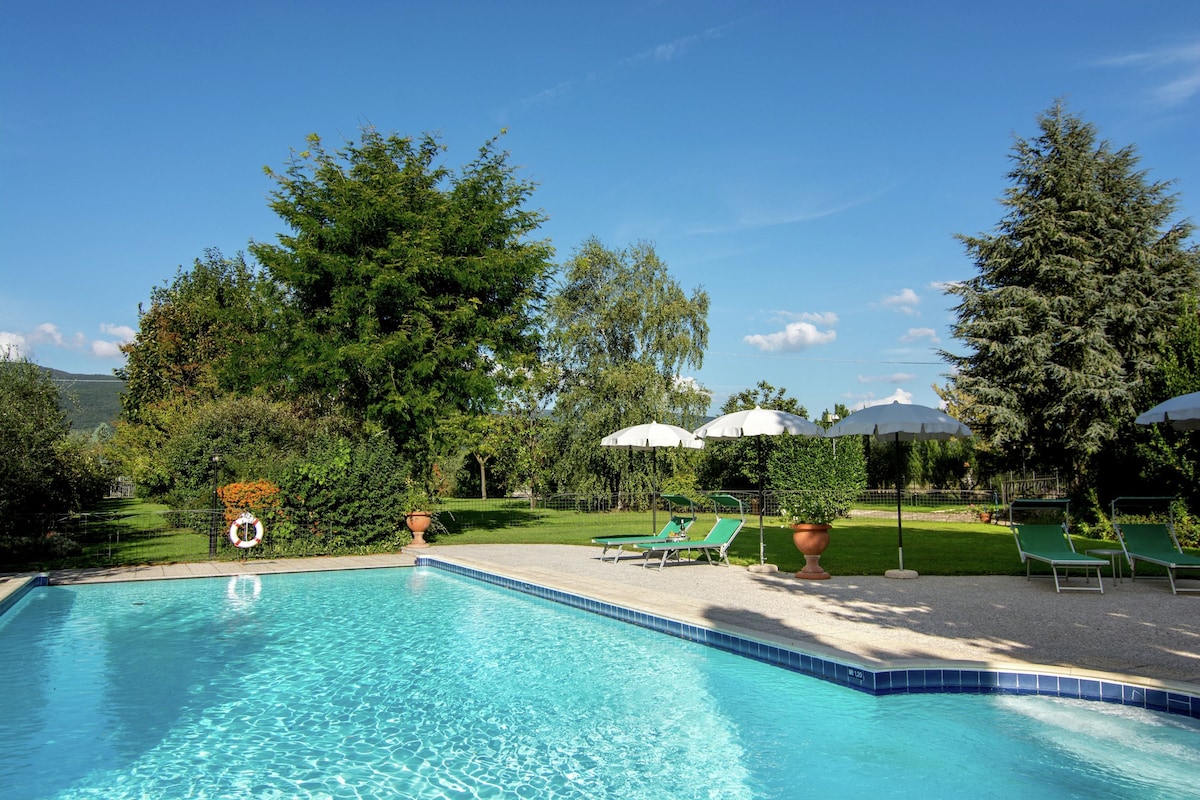 A clear blue swimming pool with lounge chairs and umbrellas, surrounded by trees and a grassy area.