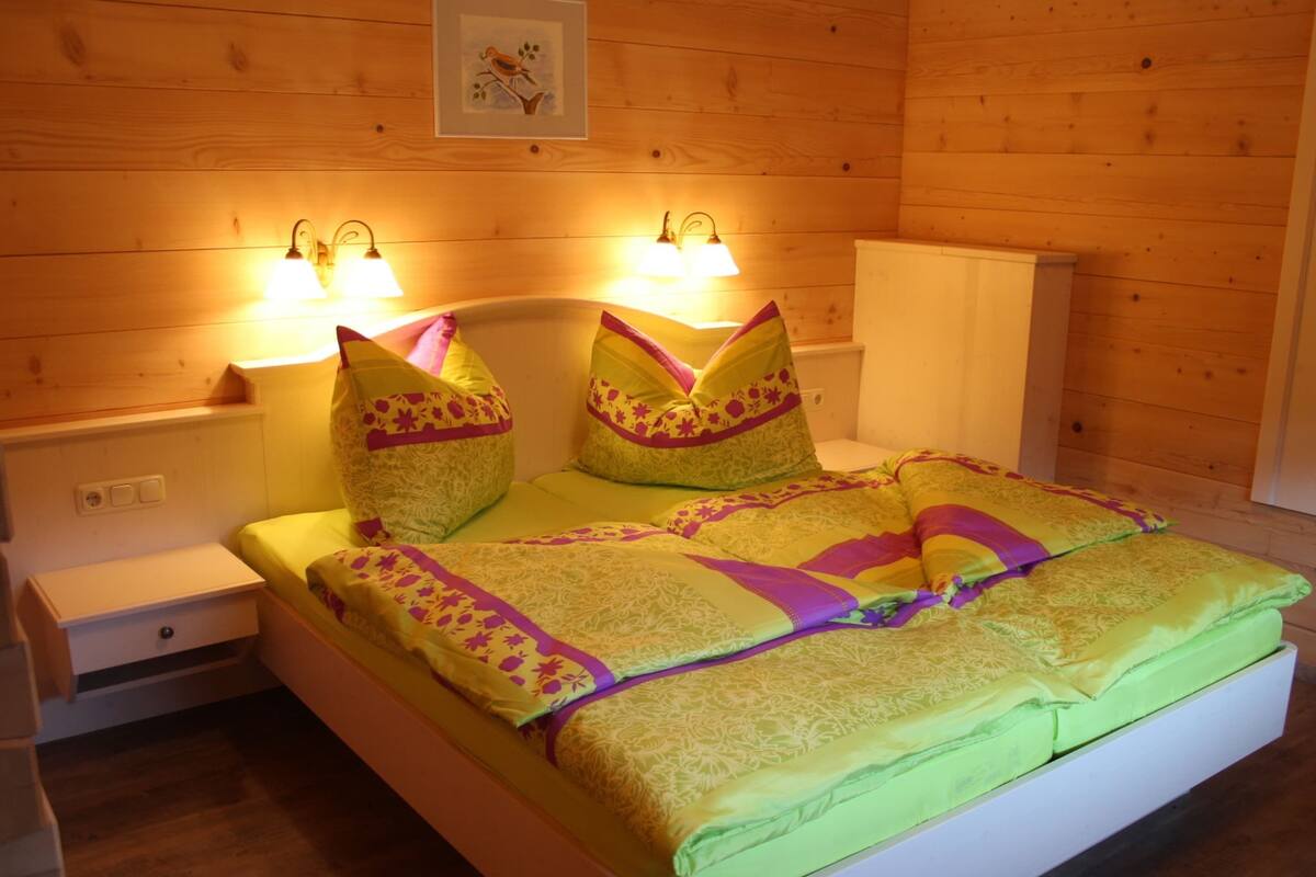 Ein Bett mit einer grünen Matratze, zwei Kissen und einer gemusterten Decke. Es gibt zwei Nachttische mit Lampen und eine kleine Schublade.
