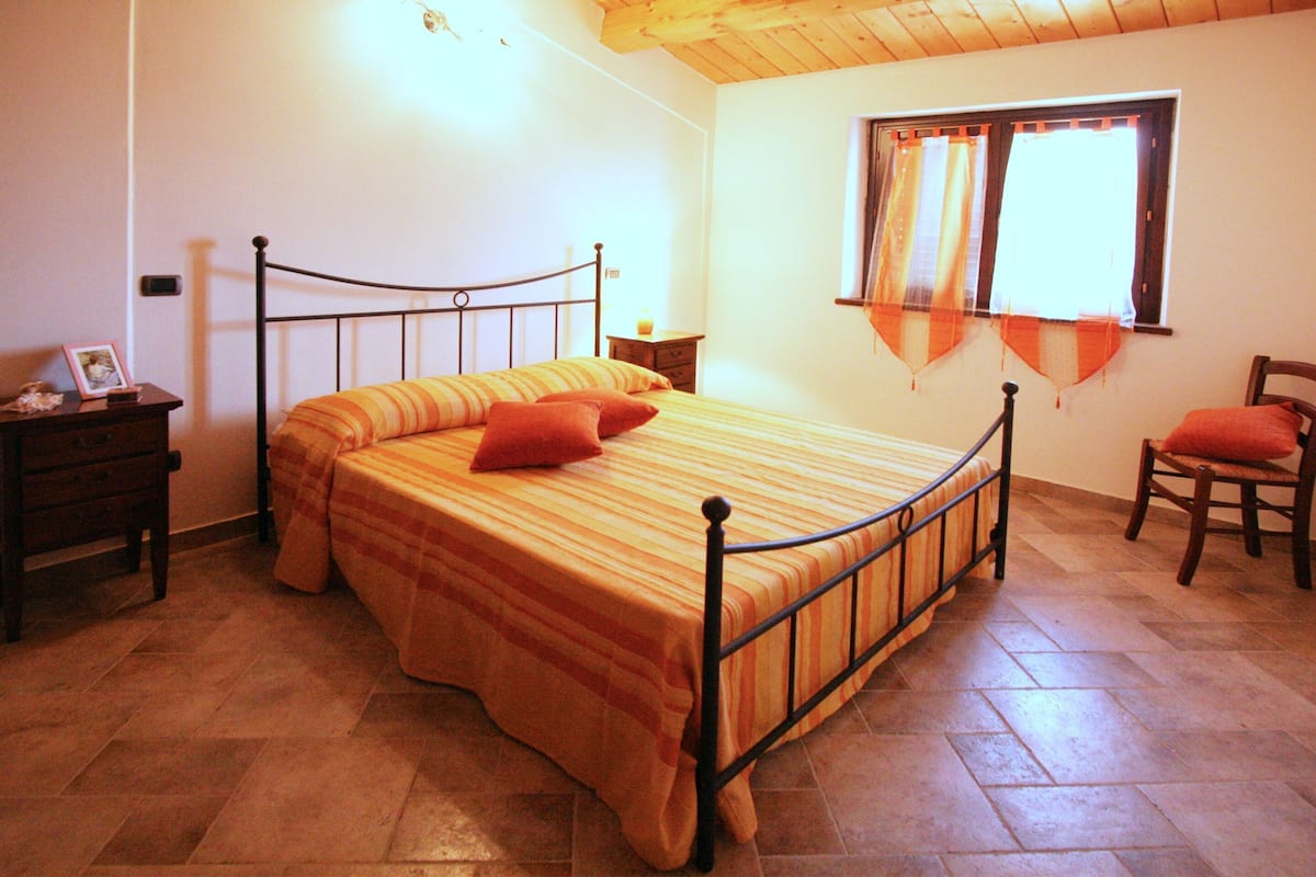 A bedroom with a bed, a nightstand, a chair, a window with curtains, and a wooden ceiling.