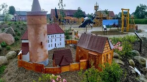 Un poble model amb una torre destacada, un edifici blanc i una casa de fusta envoltada de roques i vegetació.