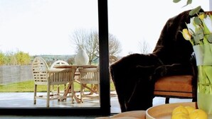 Een kamer met een groot raam, een houten tafel met stoelen, een bank en een vaas met bloemen en citroenen.