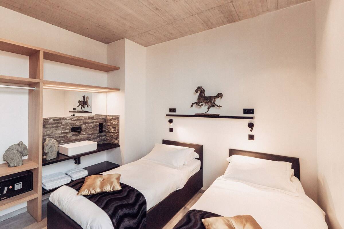 Ein modernes Hotelzimmer mit zwei Einzelbetten, einer Holzzahlendecke, einer Wandverkleidung aus Stein und einem fest montierten Waschbecken, sowie einer dekorativen Skulptur eines Pferdes.