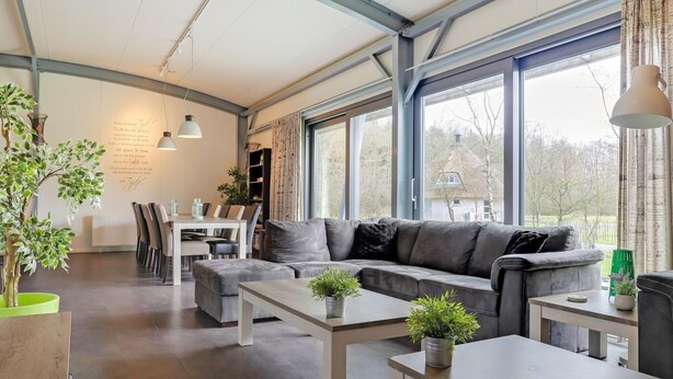 Ein modernes Wohnzimmer mit grauem Sofa, einem Esstisch mit Stühlen und großen Fenstern mit Blick ins Freie.