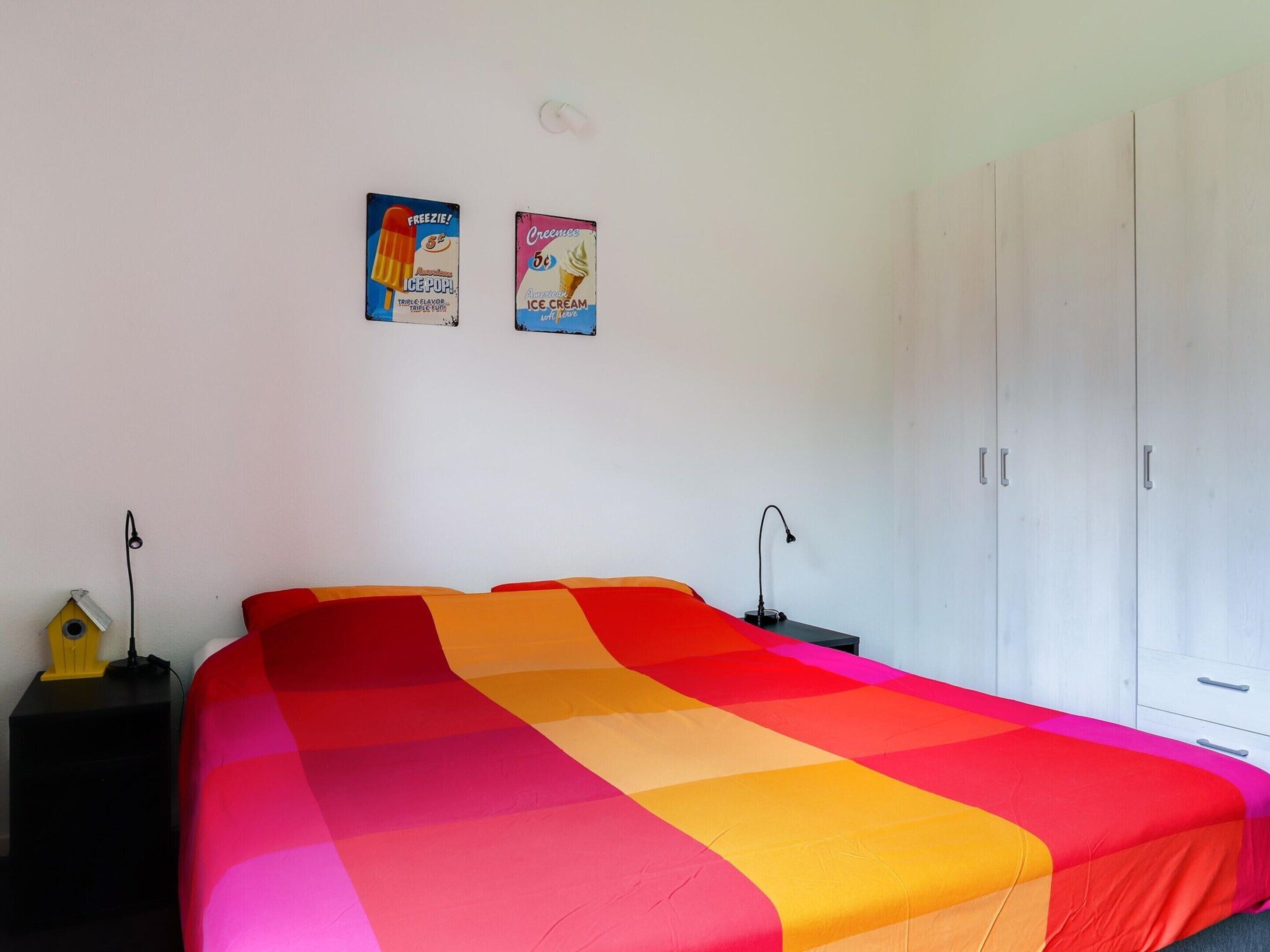 A bedroom with a bed covered in a multicolored quilt, a nightstand with a lamp, and a wardrobe.