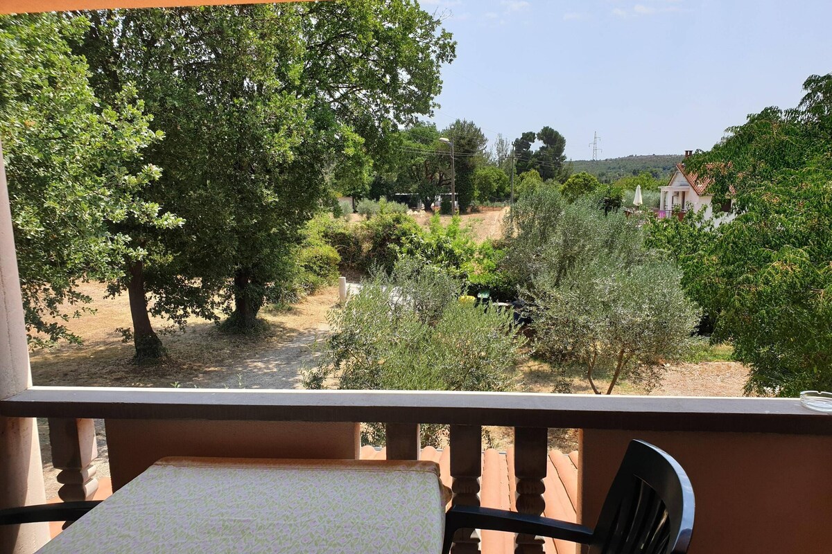 A balcony with a table and chair, overlooking a garden with trees and a distant view of a house.