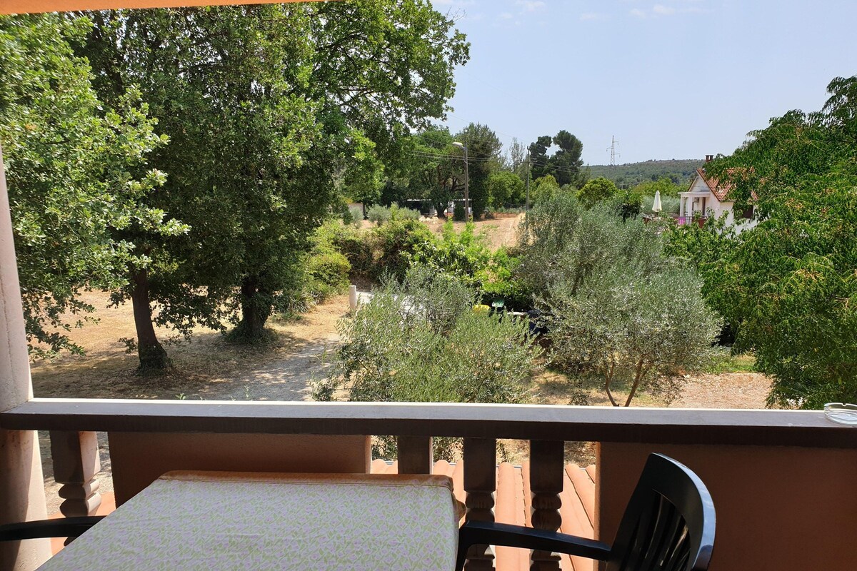A balcony with a table and chair, overlooking a garden with trees and a distant view of a house.