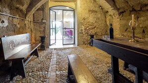 Un intérieur avec une arche en pierre menant à une pièce lumineuse, un banc en bois, une table avec une bougie et une bouteille.