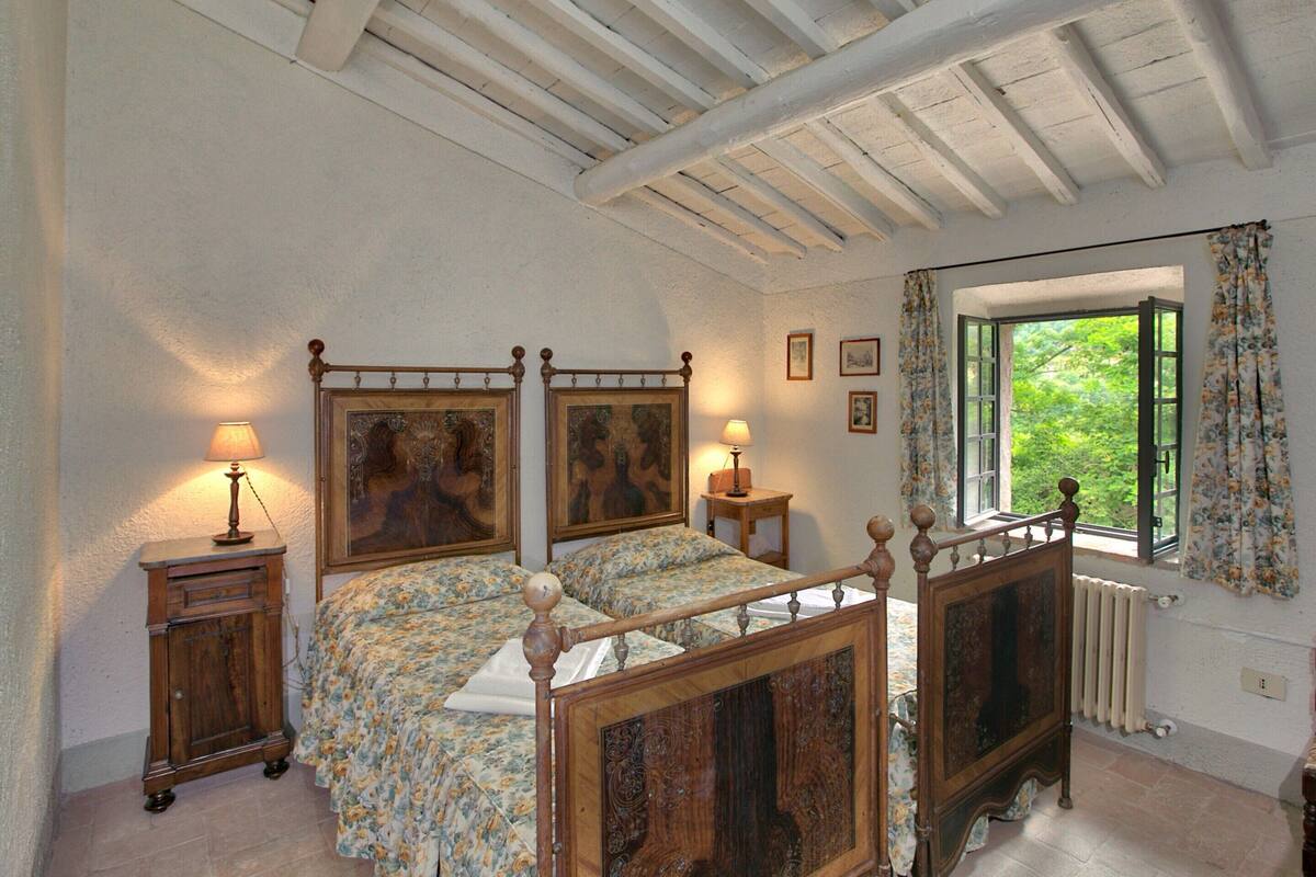 A bedroom with a wooden four-poster bed, two bedside tables with lamps, a window with a view of greenery, and a radiator.