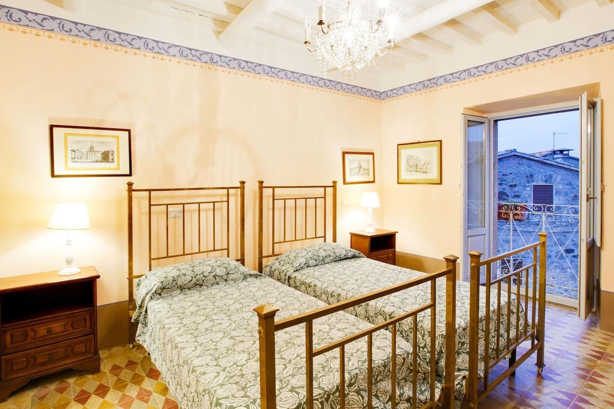A bedroom with two beds, a chandelier, and a balcony with a view of buildings.