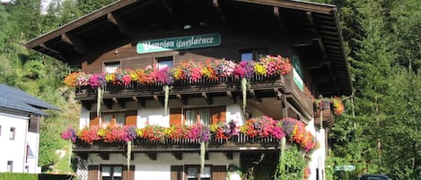 A traditional chalet with multiple balconies adorned with colorful flowers, a sign indicating it is a pension, and a car parked in front.