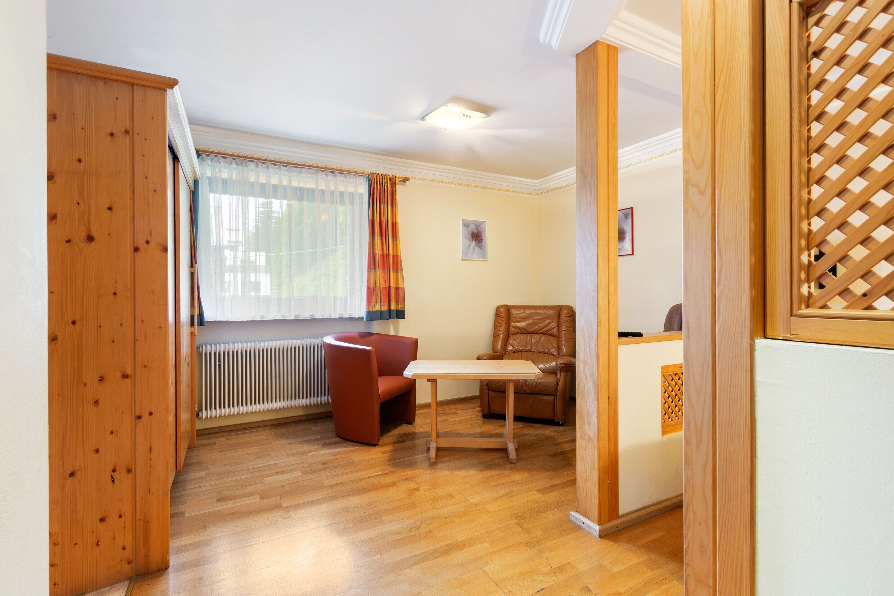 Een kamer met een houten vloer, een rode fauteuil, een klein tafeltje en een raam met gordijnen.