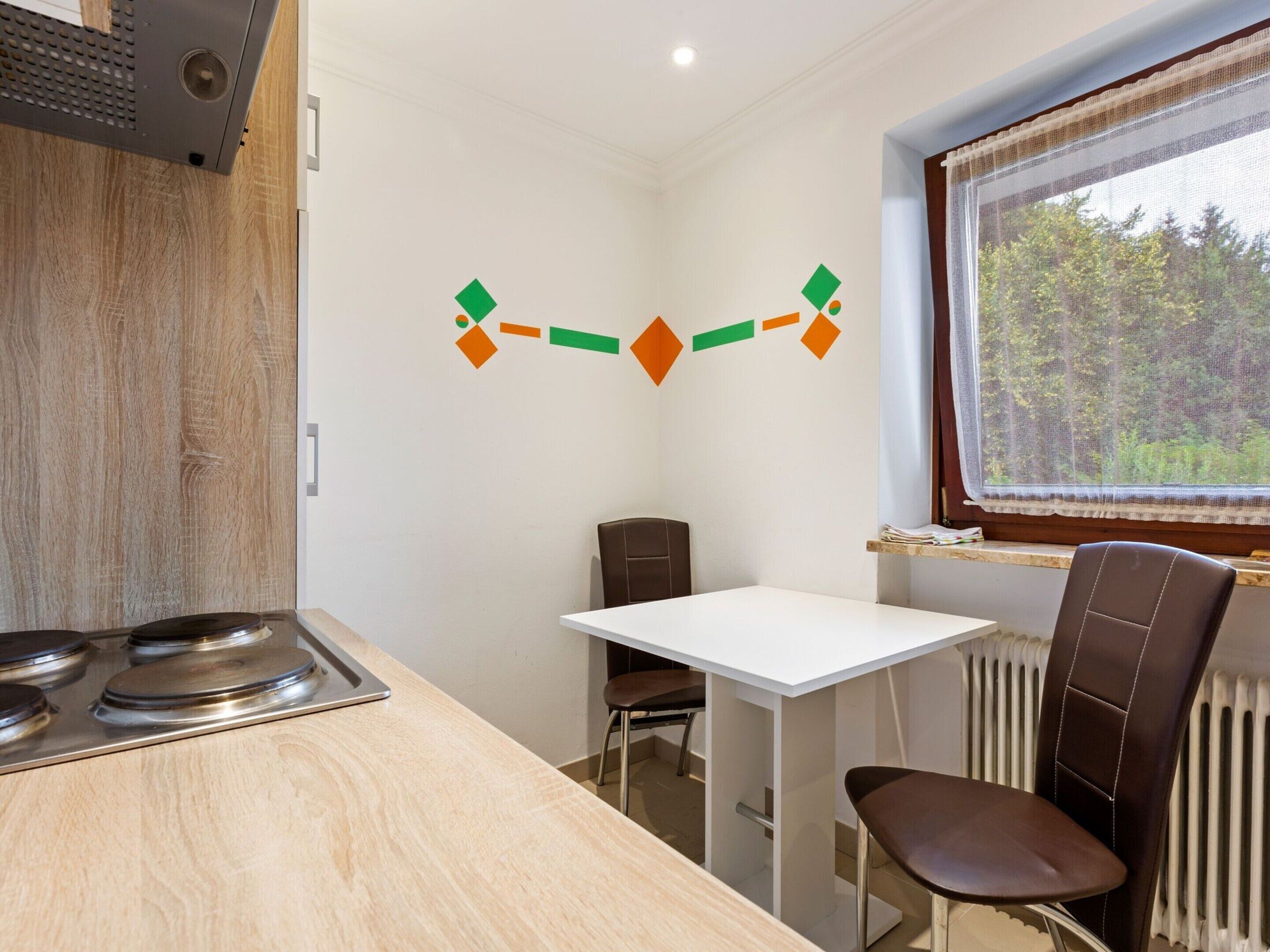 A small, modern kitchen with a white table and two chairs, a window with a view of trees, and a radiator.